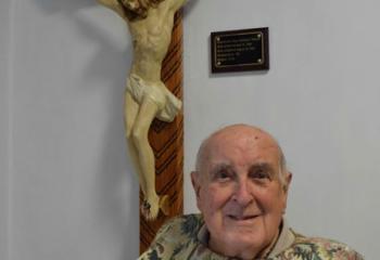 Msgr. Anthony Wassel, resident at Holy Family Manor (HFM), Bethlehem and pastor emeritus of Assumption, Sacred Heart and St. Joseph, Mahanoy City, sits beneath a cross he constructed from matchsticks and gifted to HFM. (Photo by John Simitz)