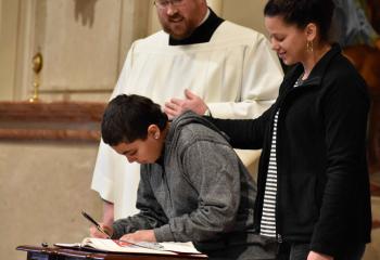 Jimmy Torres signs the Book of the Elect with the support of Anais Vega, right, and Father Allen Hoffa, administrator of St. Joseph, Summit Hill.