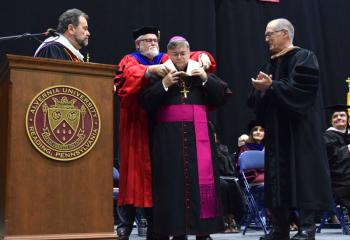 Bishop Schlert receives the hood signifying his honorary degree from Jerry Greiner. Watching are Dr. Thomas Flynn, left, and Kevin St. Cyr.