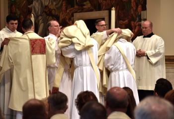 The newly ordained deacons are vested in the stole and dalmatic, which are the proper vestments for a deacon when assisting at Mass. Standing are, from left: Deacon Giuseppe Esposito, vested by Deacon Noe Ramirez de Paz; Deacon John Maria, vested by Deacon Rick Lanciano; and Deacon Zachary Wehr, vested by Deacon John Hutta. At right is Father Allen Hoffa.