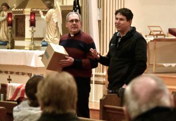 DeMatte, right, explains that Father David Loeper, left, holds a box and, not knowing the box’s content, will have to “sell” it to the faithful by answering their descriptive questions about the item. Parishioners already knew it was a roll of toilet paper when they posed questions to Father Loeper.