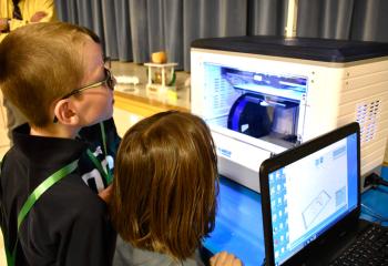 Students observe the making of a cross on a three-dimensional printer.